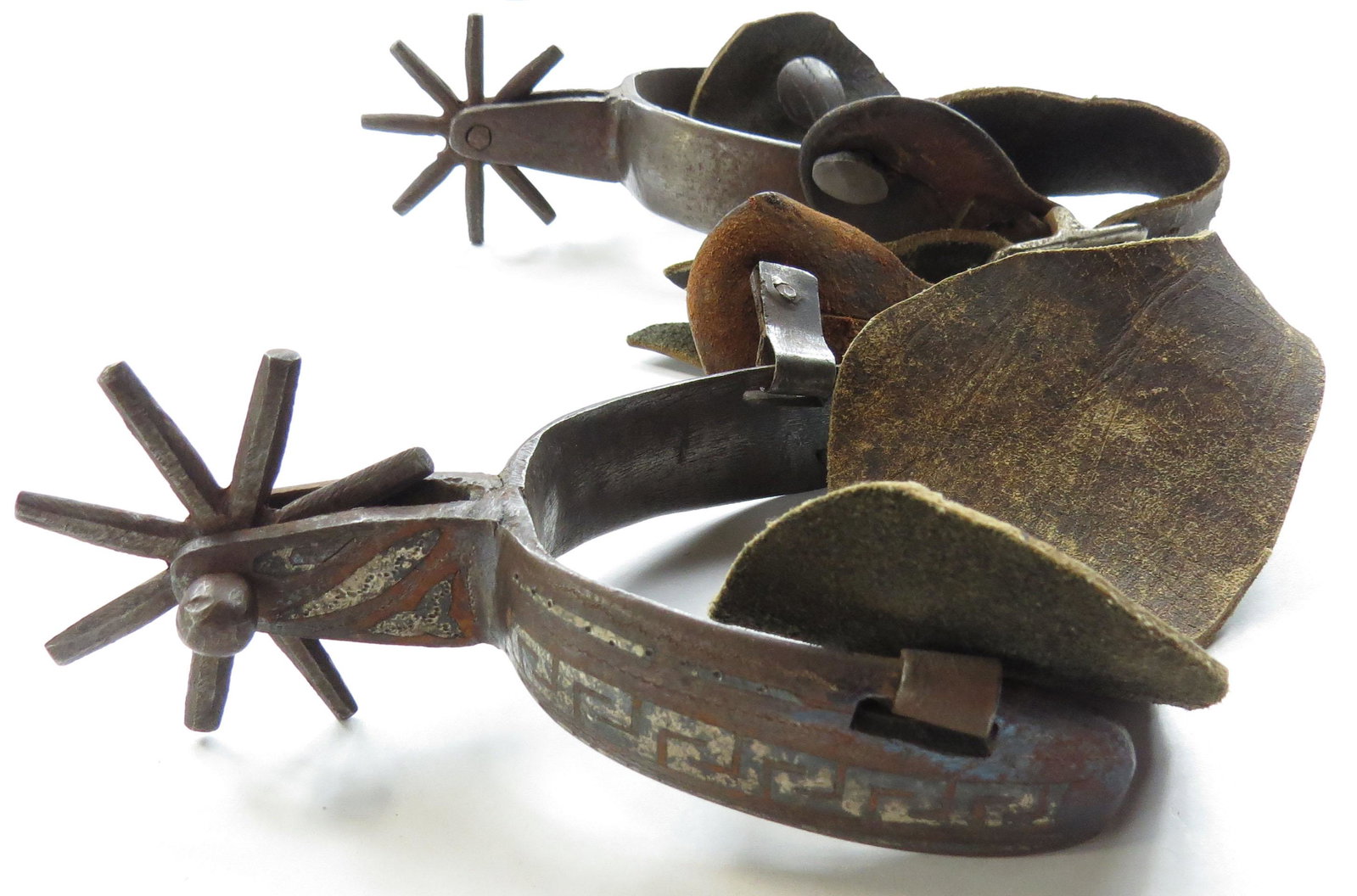A PAIR OF MEXICAN SPURS: Characteristic of older Chihuahua work, wrought entirely of steel and finely inlaid in silver with a Greek key design, with eight-pointed rowels. With older tooled straps intact. Spurs late 19th-early