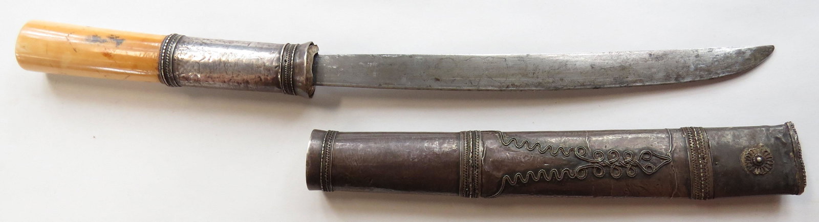 A BURMESE DHA DAGGER: The silver-mounted hilt with bands of filigree and elephant grip. Well-forged, single-edged blade. In its filigreed silver-covered wooden sheath. Latter 19th century. Overall length 34.7cm. Condition 