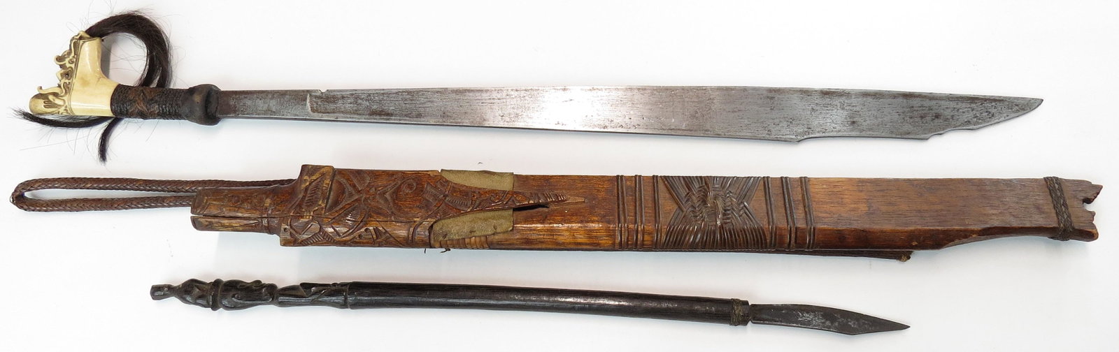 A DAYAK HEADHUNTER MANDAU SWORD: An above average example. The antler grip of fine and unusual form, well carved with aso motifs, with rattan wrapped grip. The well-wrought blade faceted, with stepped tip. In its well-carved scabbard