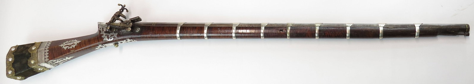 A FINE OTTOMAN SHISHANA MUSKET: A massive example stocked in finely striped hardwood of characteristic form, superbly embellished with profiled and engraved silver plaques for its entire length, signed WORK OF ASSAD MEHMET MATI. The