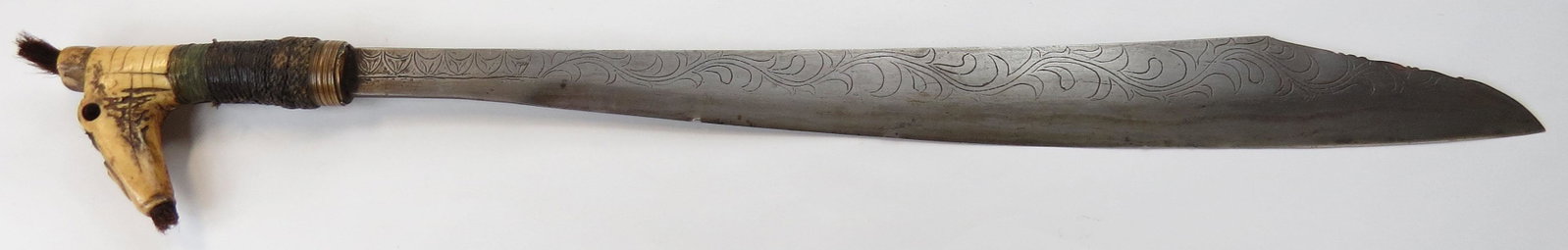 A DAYAK MANDAU SWORD: The antler grip with hair tassel. The well-wrought, chisel-edged blade showing layered construction and elaborate engraving. First half of the 20th century. Worn. Overall length 61cm. Condition III