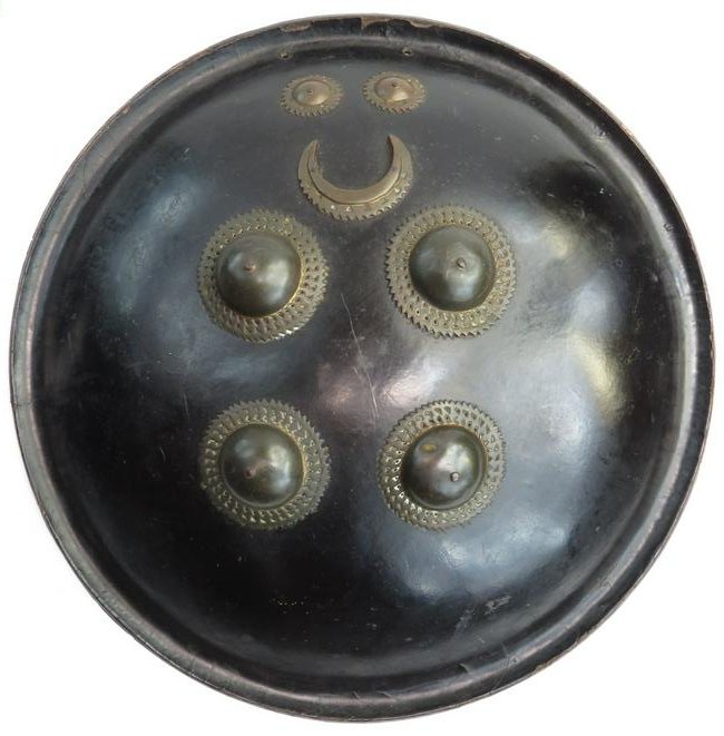 AN INDIAN DHAL SHIELD: Of circular, lightly domed form with rolled edge, formed of hardened hide, and black lacquered overall. With four pierced brass bosses a pair of suns and a crescent. Latter 18th century. Wear to lacqu