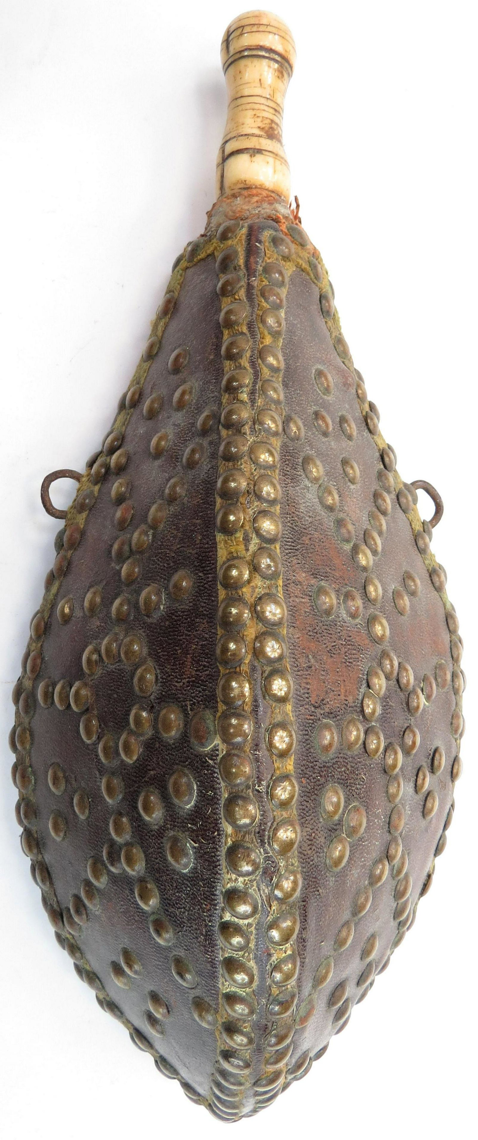 A MOROCCAN POWDER FLASK: The wooden body of characteristic lozenge form, covered entirely in leather and finely embellished with brass studs over gilt braid and turned bone spout. First half of the 19th century. Light wear. O