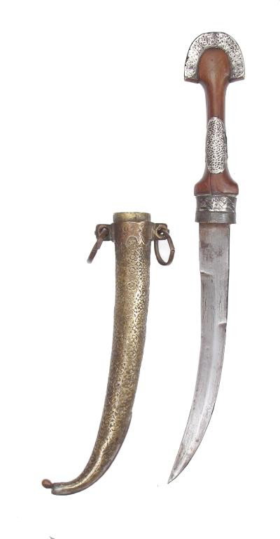 A MOROCCAN KOUMIYYAH DAGGER: Of typical form, with silver-mounted wooden grip. Blade lightly curved and double-edged, struck at the forte with maker’s marks. In its well-engraved brass scabbard with suspension rings.Early 20th 