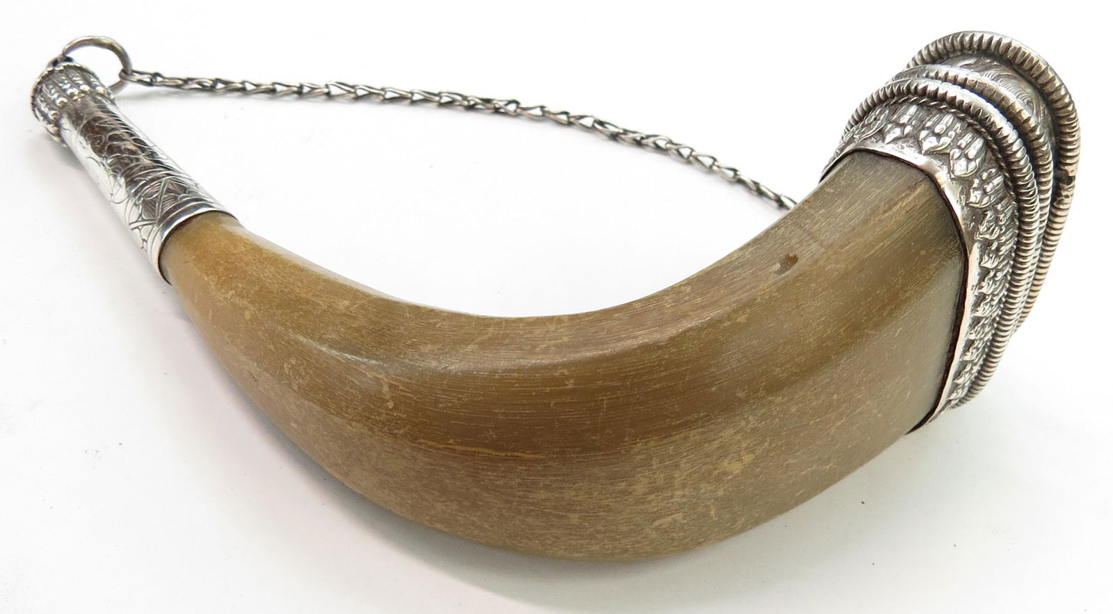 A TIBETAN POWDERHORN: The blond horn body flattened, faceted and mounted in silver, engraved in Buddhist motifs. The cap and spout retain their silver chain. Minor wear overall. First half of the 19th century or earlier. O