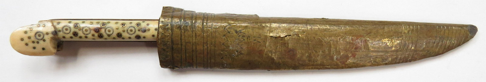 A BOSNIAN DAGGER: The eared grips of bone, well- inlaid with brass. Broad, single-edged blade with maker’s mark at left forte. Sheath of wood covered in brass with incised lines. 19th century. Minor losses to brass.