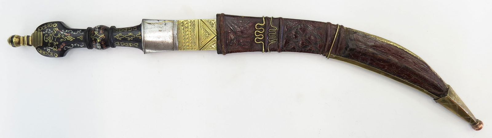 A FINE UNUSUAL TUAREG DAGGER: The well-formed hardwood grip elaborately inlaid with brass and silver wire, the curved, double-edged blade with silver ferrule and engraved brass bolster. In its tooled leather scabbard with brass mo