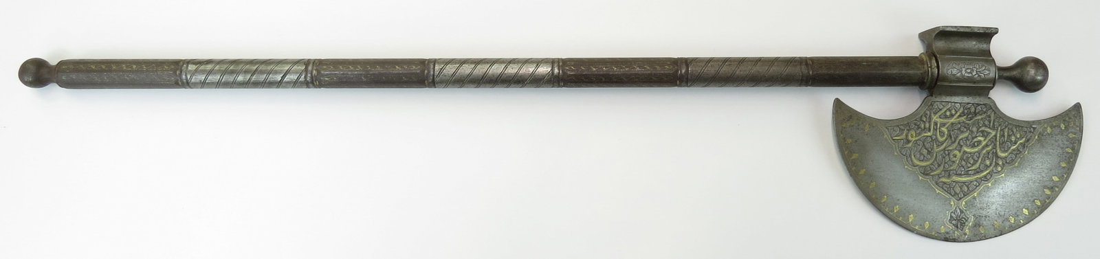 A PERSIAN BATTLE AXE: Wrought entirely of steel, the head of crescent form with flared hammer peen, with finely chiseled panels containing Qur?anic inscriptions inlaid in gold. On its engraved steel haft. 19th century. Wor