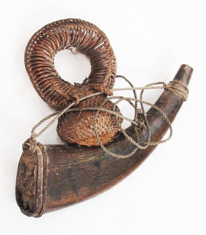 A RARE TIMORESE POWDERHORN AND BALL CONTAINER: The horn with good age patina and original cord, retaining its circular woven rattan ball- holder, the opening closed with wool also used for patching. Mid-19th century.Overall length 21.9 cm. Conditi