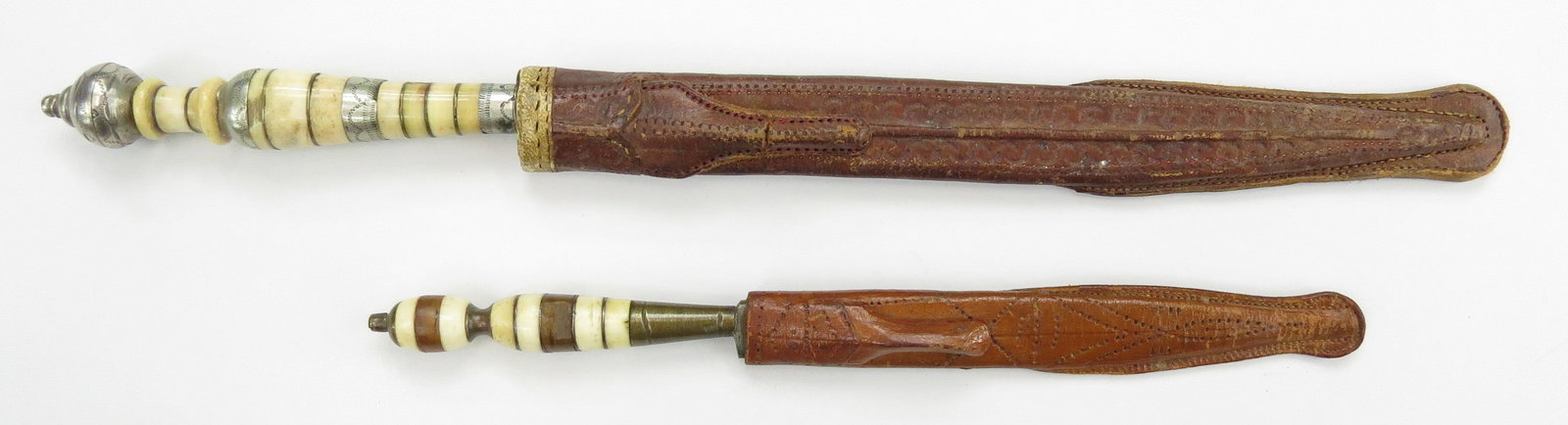 TWO BRAZILIAN FACON DAGGERS: Of characteristic form, with segmented bone and silver grip and spherical pommel on the larger. In their tooled leather sheaths. Late 19th-20th c. Worn. Overall length of the largest 31.2cm/ 12 1/4 in