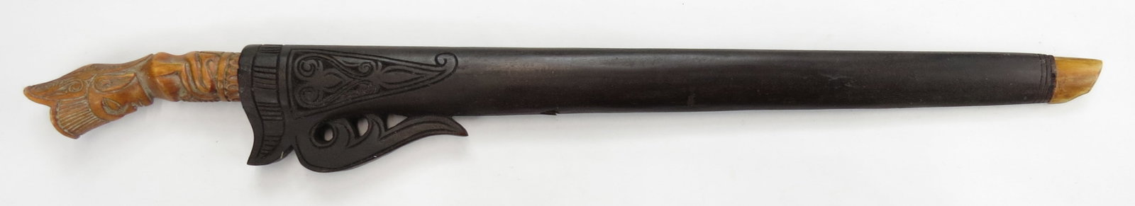 A SUMBAWAN SWORD: The carved bone handle in the form of a seated deity, the straight, single-edged and fullered blade chiseled at the forte. In its hardwood scabbard with horn tip. 19th-20th century. Worn. Overall leng