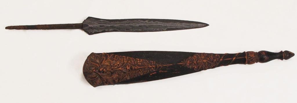 AN INDONESIAN TOMBAK SPEARHEAD: Of characteristic form, expanded at the forte and midsection, and forged of dramatically-patterned pamor, with a lengthy spike tang. The hardwood scabbard finely carved with raised panels of scrolling