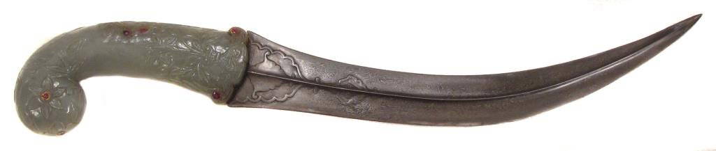 A FINE MUGHAL KHANJAR DAGGER: The grip carved of a single section of green jade, with flared, trilobate guard, tapered grip and rolled pommel, carved overall with flowering lotuses and embellished with rubies set in gold in kundan