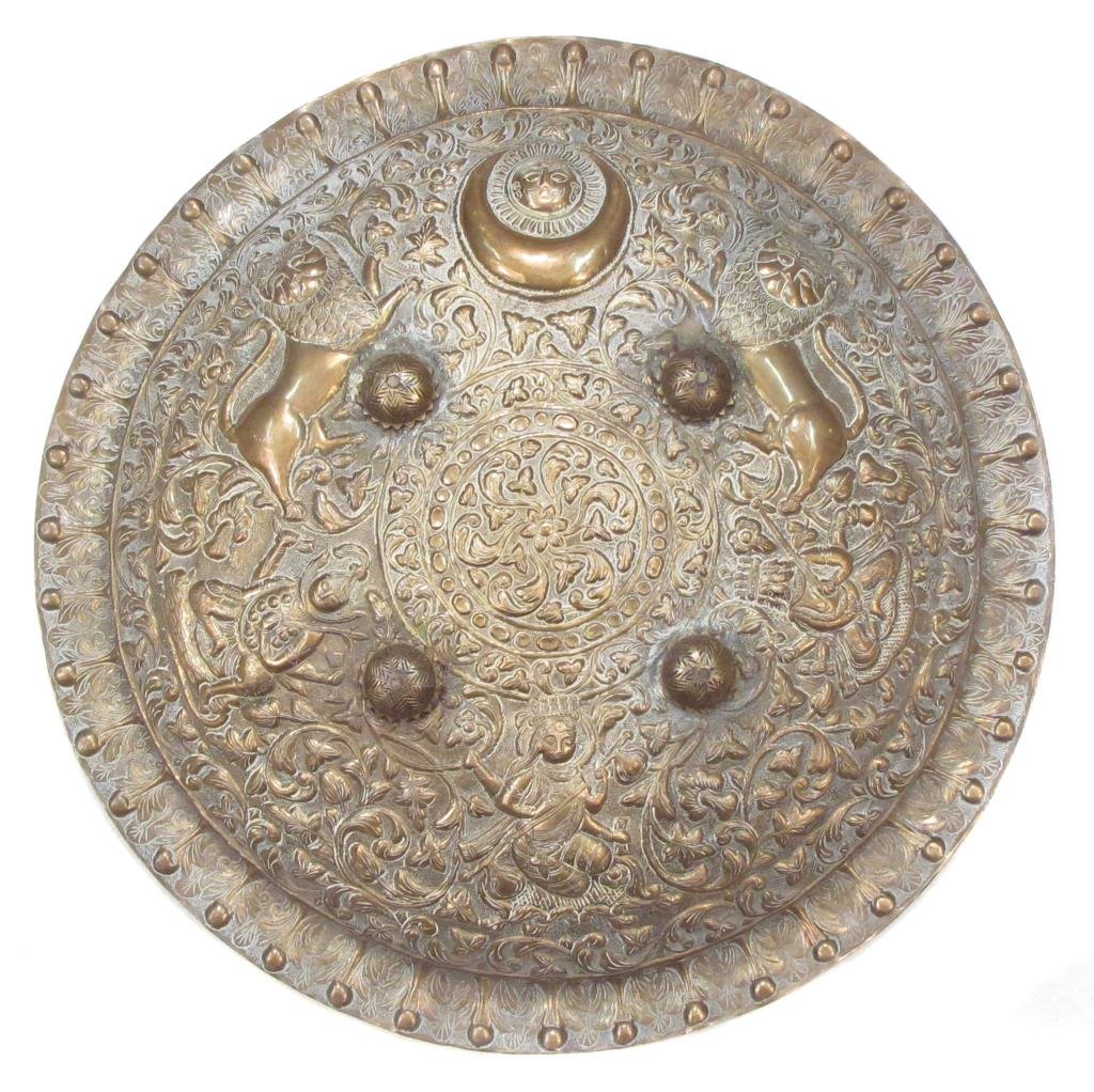AN INDIAN DHAL SHIELD: A large, highly embellished example. Handwrought of brass, and finely embossed and chased overall with lions and Indian god warriors all amongst foliage, surmounted by the Sun God, Surya. With applied