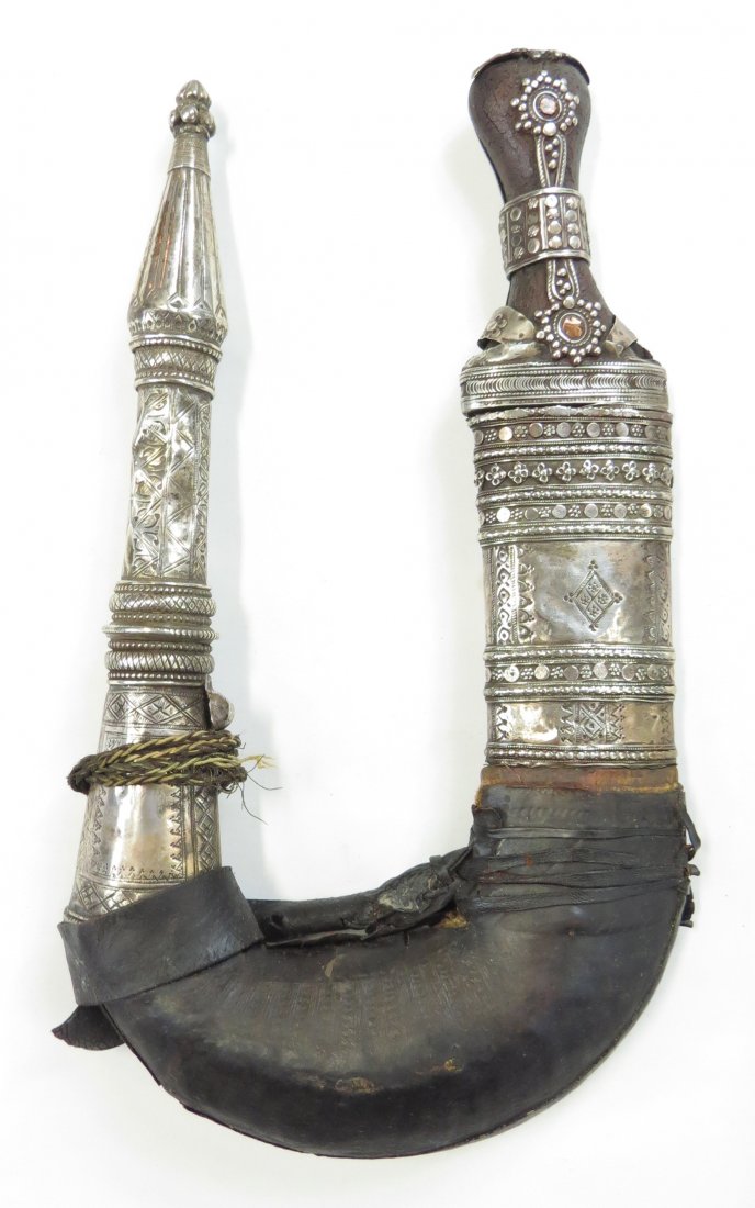 AN ARAB KHANJAR DAGGER: From Hadhramaut, known as a gusbi. The horn grip elaborately mounted in filigreed and engraved silver, the broad blade wrought with central ridge. In its leather-covered wooden scabbard with large sil