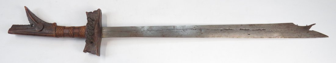 A MORO KAMPILAN SWORD: The hardwood hilt of typical form, exceptionally well-carved overall, with iron guard and rattan grip-wrap intact. The long, single-edged blade expanding toward the profiled and pierced tip. Late 19th
