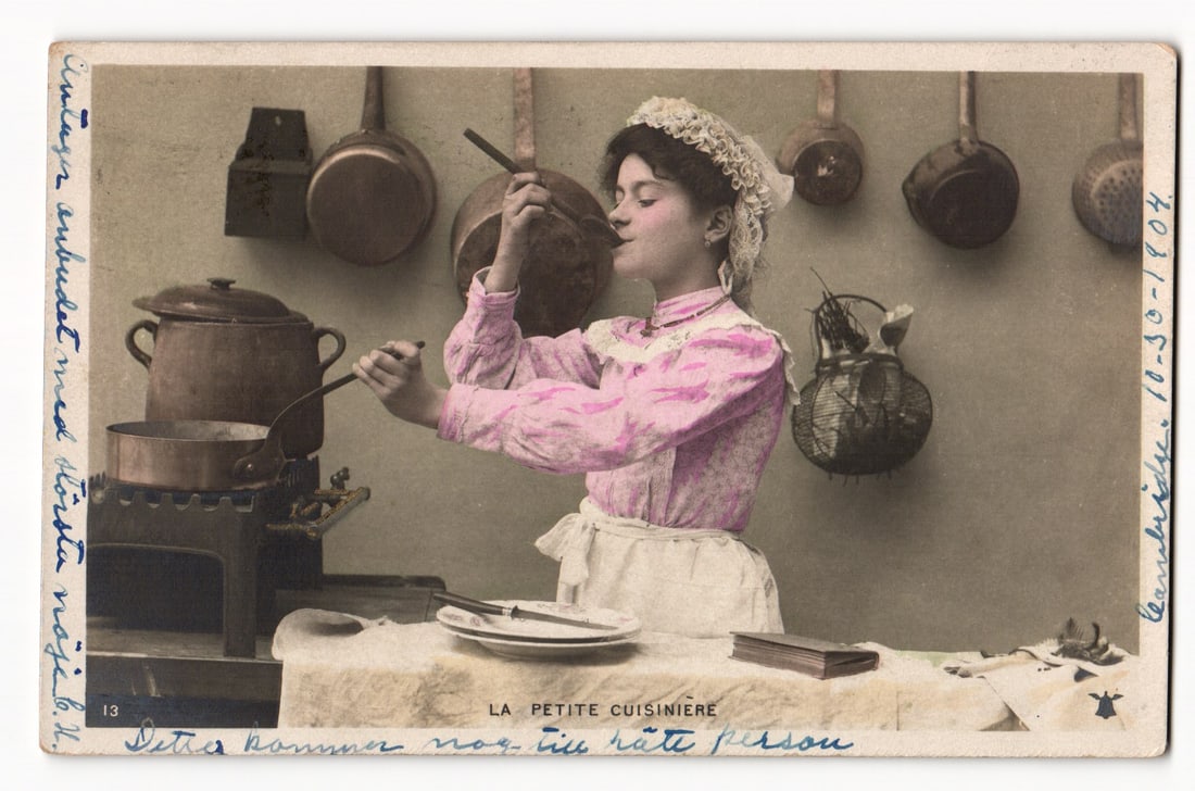 La Petite Cuisinière: Young Woman in Pink Blouse Cooking at Stove, Early 20th Century Postcard: The front of the postcard features a tinted photograph titled "LA PETITE CUISINIÈRE". The image depicts a young woman with dark hair styled under a white lace cap, wearing a pink patterned blouse wit