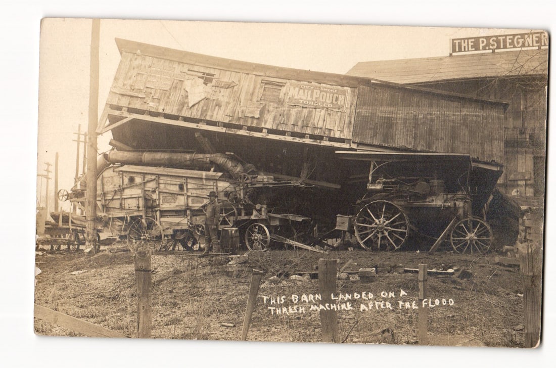 RPPC of Barn Collapsed on Threshing Machine After Flood, Mail Pouch Tobacco Advertisement Visible: The postcard front features a sepia-toned photograph depicting the aftermath of a destructive event, likely a flood as indicated by handwritten text. The primary subject is a large wooden barn structu