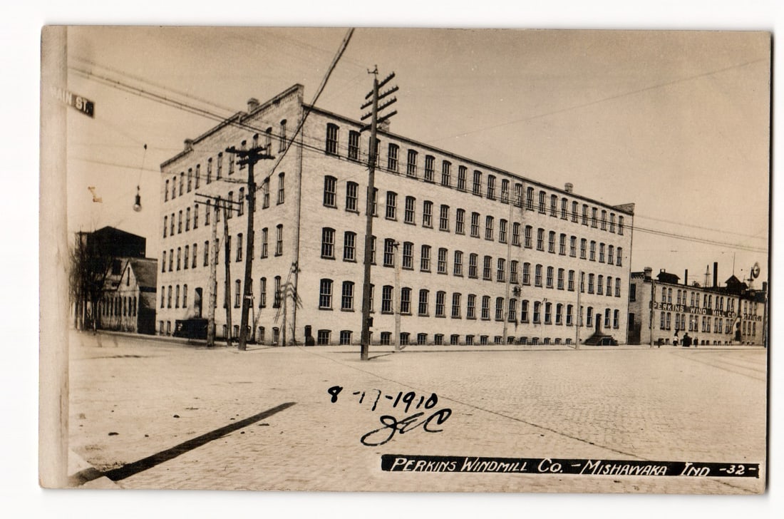 Perkins Windmill Co. Building, Mishawaka, Ind., Street View with Cobblestones, Dated 1910: The postcard presents a sepia-toned Real Photo Postcard (RPPC) view of the Perkins Windmill Co. in Mishawaka, Indiana. The primary subject is a large, multi-story, light-colored brick industrial build