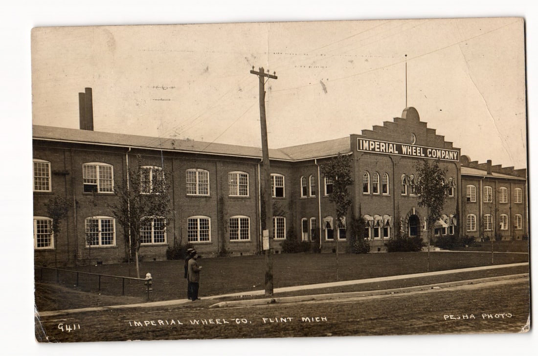 Imperial Wheel Company Industrial Building Exterior, Flint, Michigan; Two Figures, c. 1912 RPPC: The front of the postcard displays a sepia-toned photograph of a large industrial building. The structure is identified by prominent signage as the "IMPERIAL WHEEL COMPANY". It is a multi-story brick
