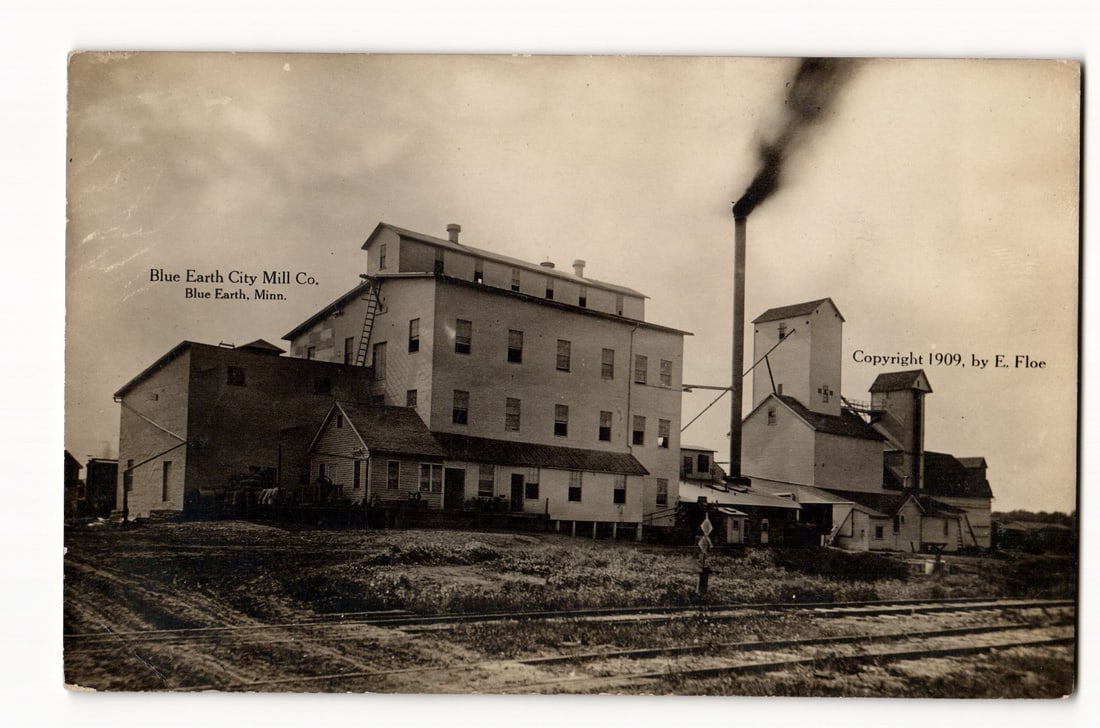 Blue Earth City Mill Co. Industrial Buildings, Railroad Tracks, Blue Earth, Minn., c. 1909 RPPC: The postcard front features a sepia-toned photograph of the Blue Earth City Mill Co. in Blue Earth, Minnesota. The image is dominated by a large, multi-story mill building, which appears to be the cen
