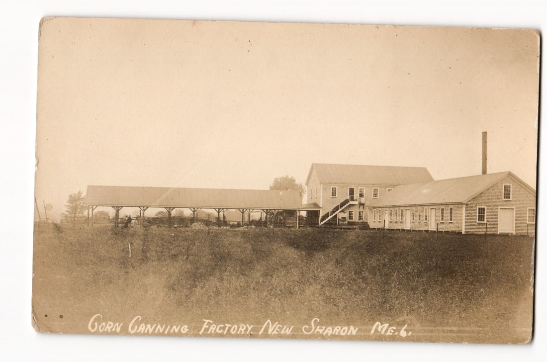 Photographic View of the Corn Canning Factory Buildings and Grounds, New Sharon, Maine, c. 1912: The postcard presents a sepia-toned photographic image, identified as a Real Photo Postcard (RPPC), depicting the Corn Canning Factory in New Sharon, Maine. The composition is an eye-level, medium-lon