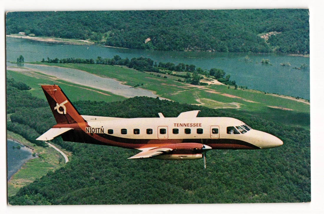 Tennessee Airways "Bandeirante" Prop-Jet N101TN in Flight, Aerial View of Forested Terrain: The postcard front displays a color photograph capturing an aerial view of a Tennessee Airways aircraft in mid-flight. The aircraft, identified by its markings and design as an Embraer EMB 110 Bandeir