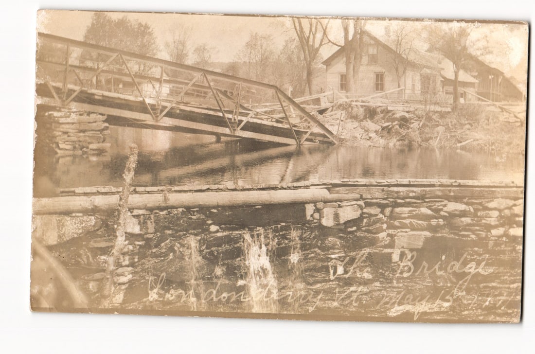 Damaged Metal Truss Bridge, Londonderry, Vermont, Aftermath of Event, May 15, 1911 RPPC: Front: The postcard features a sepia-toned photograph. The primary subject is a metal truss bridge, partially collapsed and submerged in a river. The river flows towards the viewer, with a low, rough-