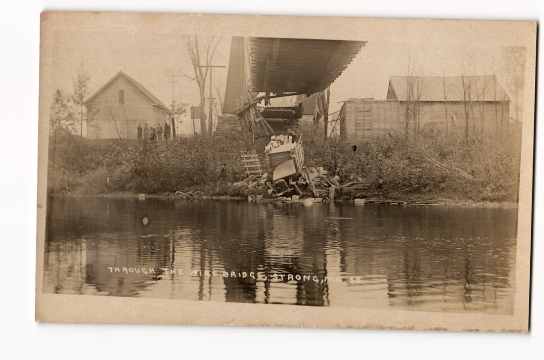 Real Photo Postcard: Vehicle Fallen Through Collapsed Wire Bridge, Strong, Maine Accident Scene: The front features a sepia-toned photograph depicting the aftermath of a structural failure where a vehicle has plunged through a collapsed bridge. The perspective is from the water, looking towards t