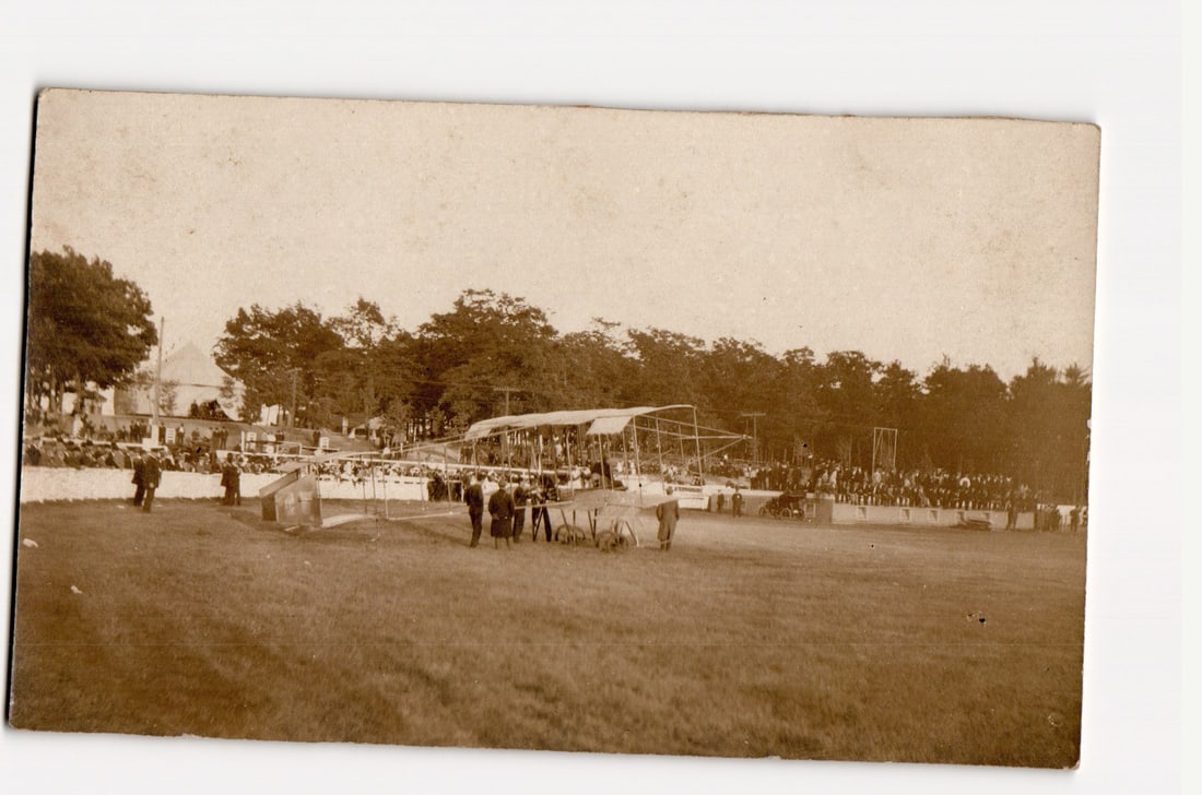 Early 20th Century Biplane Aircraft on Grassy Field, Large Crowd of Spectators, Aviation Meet: The front of the postcard displays a sepia-toned photograph. The central subject is an early model biplane aircraft resting on a large, open grassy field. A significant crowd of spectators is gathered