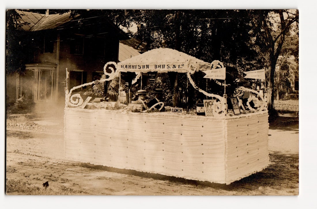Harrison Bros. & Co. Parade Float or Stand, Lebanon, New Hampshire Early 20th C. Photo Postcard: A sepia-toned, outdoor photograph captures a parade float positioned on a dirt road. The float, extensively decorated with a white, tufted material covering its sides, features a canopy. Atop the floa