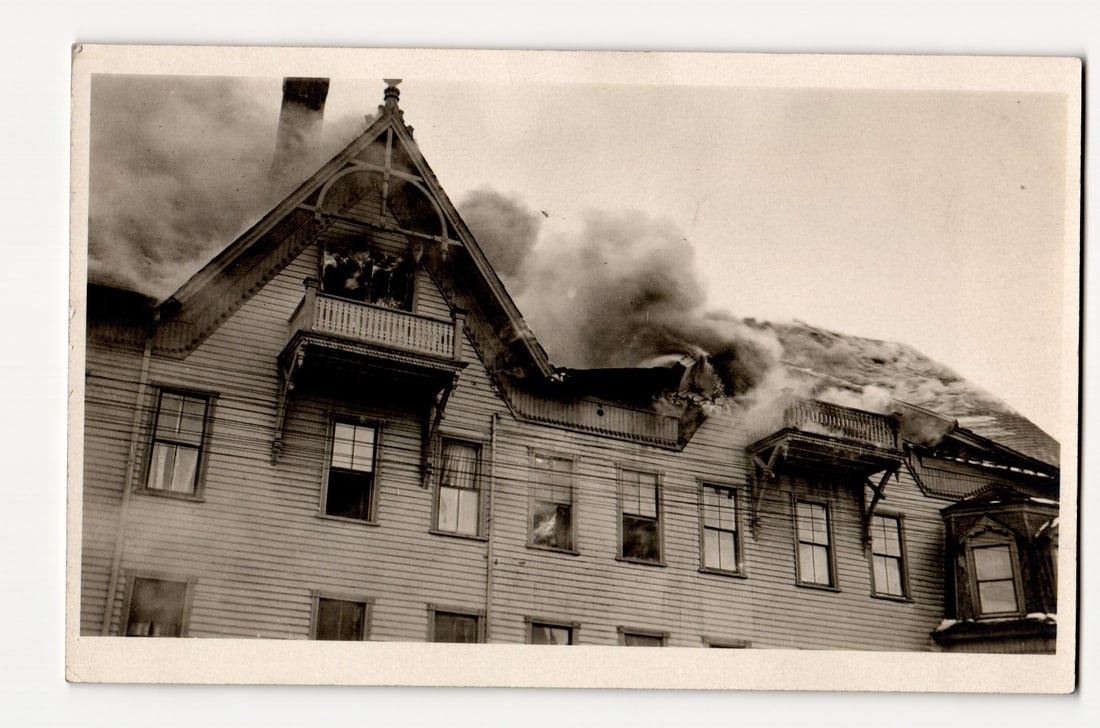 Sherburne, N.Y. Building Fire, Smoke Billowing from Roof, Figures on Balcony, RPPC Postcard: A black and white photograph captures a multi-story wooden building, characterized by clapboard siding and multiple gables, from which dense smoke is emanating, primarily from the roofline and upper l