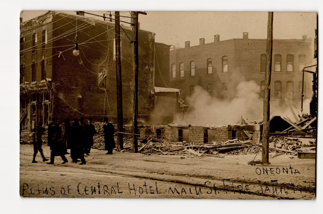 Oneonta, NY: Ruins of Central Hotel After Main Street Fire, January 1, 1911 Real Photo Postcard: A sepia-toned Real Photo Postcard (RPPC). The image depicts the aftermath of a fire. In the foreground, a street with what appear to be trolley tracks is visible, and a group of at least seven figures
