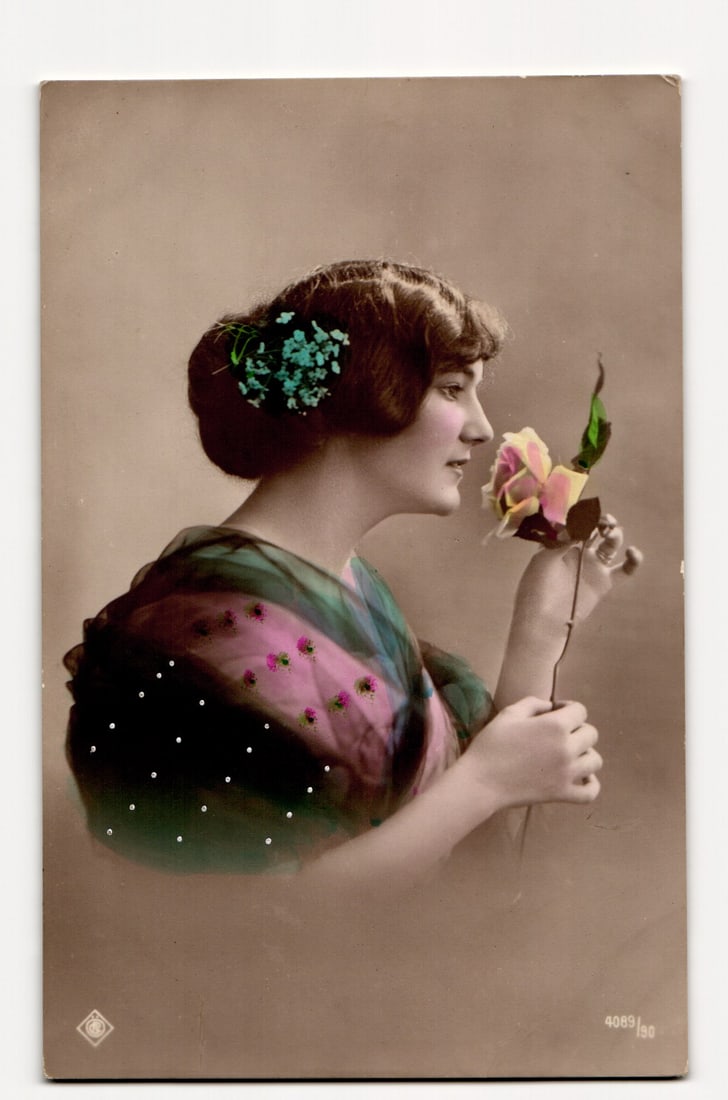 Hand-Colored Photographic Postcard: Woman in Profile Smelling Rose, Teal Flowers in Hair Detail: The front of the postcard features a hand-colored photographic portrait of a woman in profile, facing right. She has dark hair styled in an updo, adorned with small, teal-colored flowers. The woman is