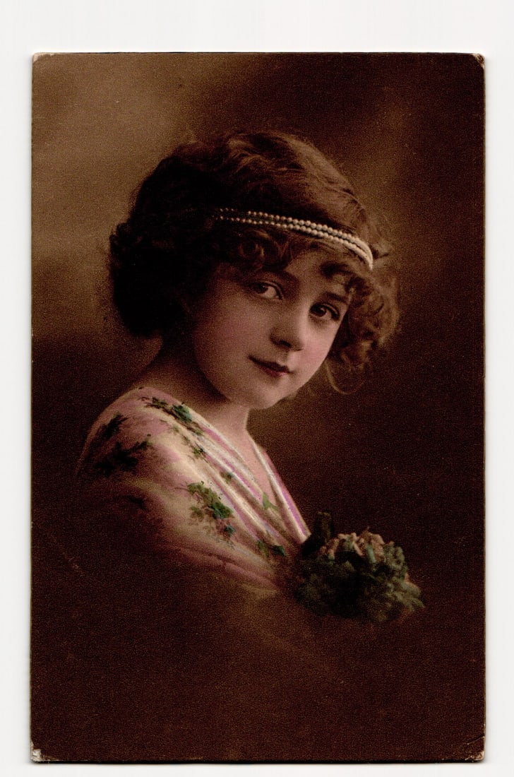 Hand-Tinted RPPC Postcard of Young Girl with Pearl Headband, Voro-Gravure Serie 85, Germany: A sepia-toned, hand-tinted photographic portrait depicts a young girl with dark, curly hair adorned with a pearl-like headband. She is shown from the chest up, her body slightly angled, looking direct