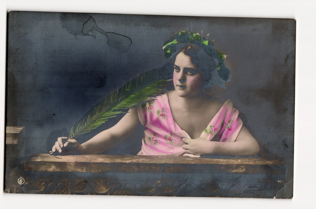 INNOCENCE.? Woman with Palm Frond, Austrian Colorized Photograph Postcard, Early 20th Century: A colorized photographic portrait depicts a young woman positioned behind a wooden ledge with a textured lower section. She has dark hair adorned with a green wreath and wears a light pink, sleeveless