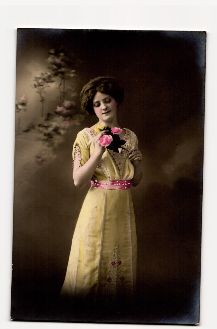Woman in Yellow Dress Holding Pink Roses, Hand-Tinted Early 20th Century Real Photo Postcard: A full-length portrait of a young woman, standing and angled slightly right, gazing down at a small bouquet of three pink roses held in both hands. She has dark, upswept hair. The woman wears a pale y