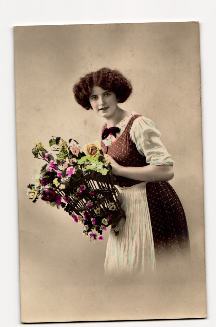 Hand-Tinted RPPC Postcard: Woman in Folk Dress with Large Floral Basket, Polish Text on Verso: A hand-tinted photographic postcard depicting a young woman with reddish-brown, curly hair. She is attired in a dark, polka-dotted bodice over a white blouse with puffed sleeves, and a light-colored a