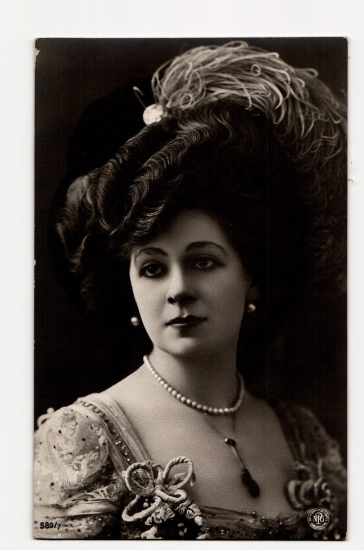 RPPC Postcard Portrait of an Elegant Woman with Feathered Hat and Pearls, NRG Logo, No. 589/7: A sepia-toned photographic portrait of a woman from the shoulders up, angled slightly to her right, with her gaze directed slightly to the viewer's left. She wears a large, dark hat adorned with abund