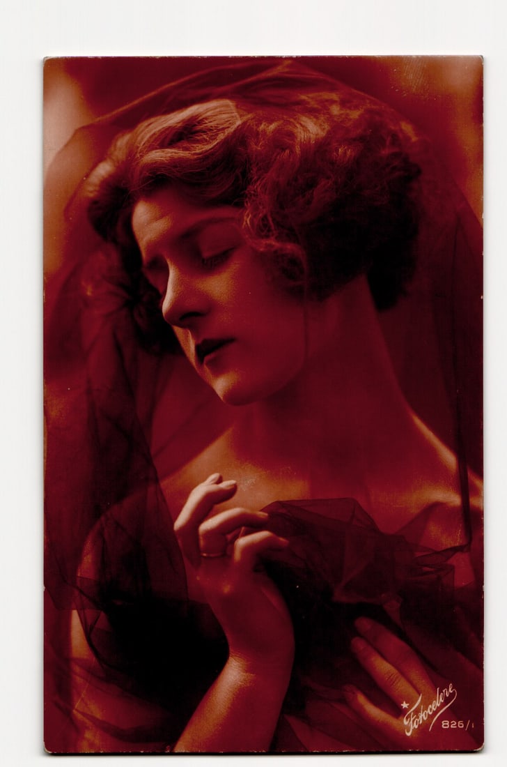 Fotocelere B26/1 Tinted Portrait Postcard of Woman with Sheer Veil, Made in Italy, circa 1920s: A tinted photographic postcard featuring a close-up portrait of a woman with reddish, curly hair. Her eyes are closed as she gazes downwards and to her left. She is enveloped in a dark, sheer fabric,