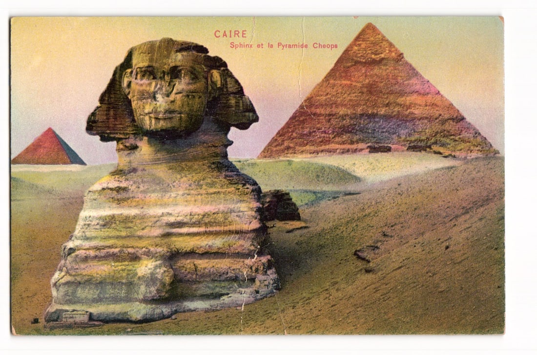 Cairo, Egypt: The Sphinx with the Pyramid of Cheops in the background, colorized postcard.: A colorized image shows the Sphinx in the left foreground, facing three-quarters to the right. Significant weathering is visible on its facial features and body. To the right and further back, the Pyr