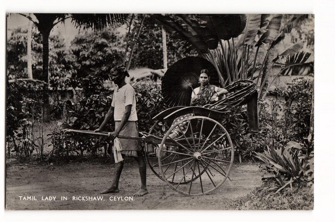 Photograph Postcard: Tamil Lady in Rickshaw, Ceylon. Early 20th C. Publ. Plâté, Ltd.: A black and white photograph depicts a woman, identified by printed text on the front as a "Tamil Lady," seated in a pulled rickshaw. The woman is dressed in a patterned sari and holds a dark parasol.