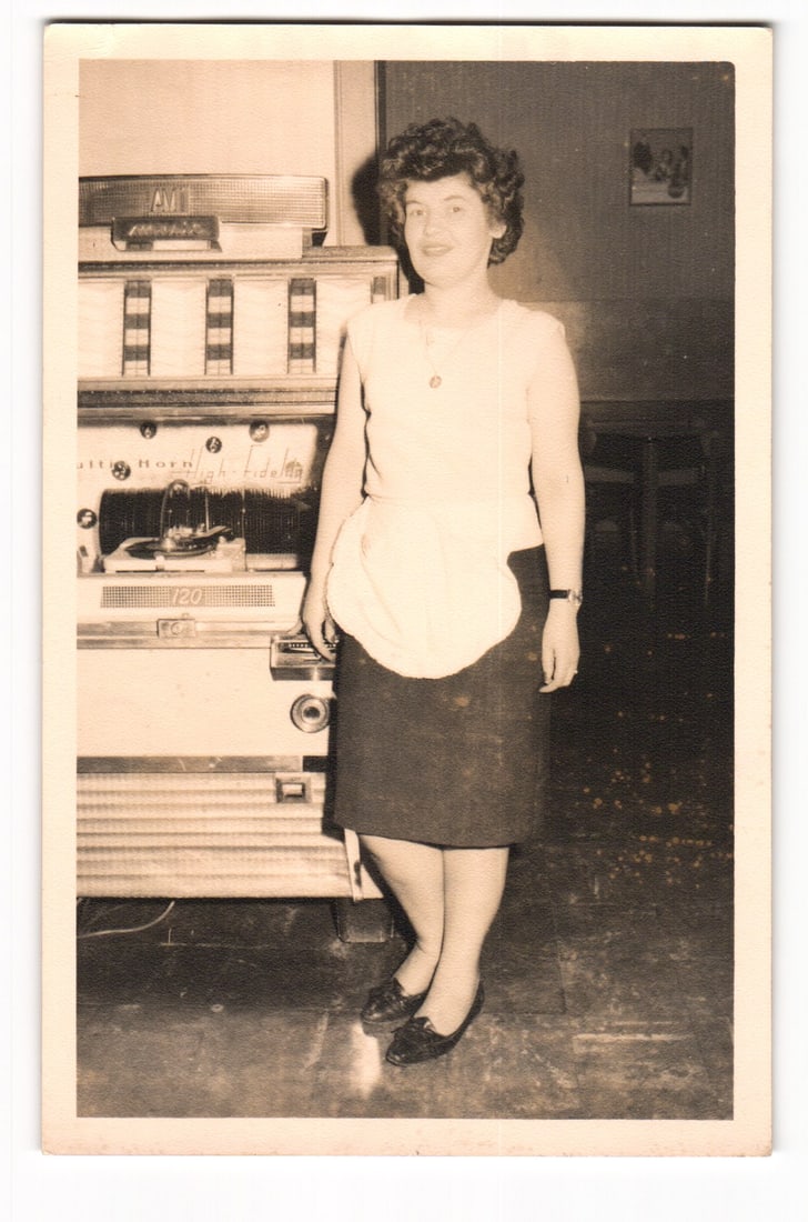 RPPC Postcard Woman Waitress Beside AMI G-120 Jukebox Germany c. 1950s Foto-Gust Lieblos: This Real Photo Postcard (RPPC) presents a sepia-toned, full-length photographic portrait. The central subject is a woman with dark, curly hair styled away from her face. She is attired in a light-col