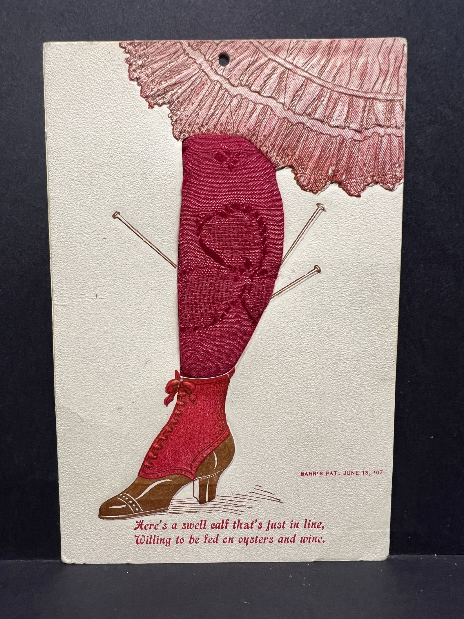 Humorous Fabric Appliqué Postcard, Woman's Leg & Stocking, Barr's Patent 1907, USA Origin: This postcard features a central illustration of a woman's lower leg and foot against a textured, off-white paper background. At the top, a portion of an embossed, frilled pink skirt hem is depicted.