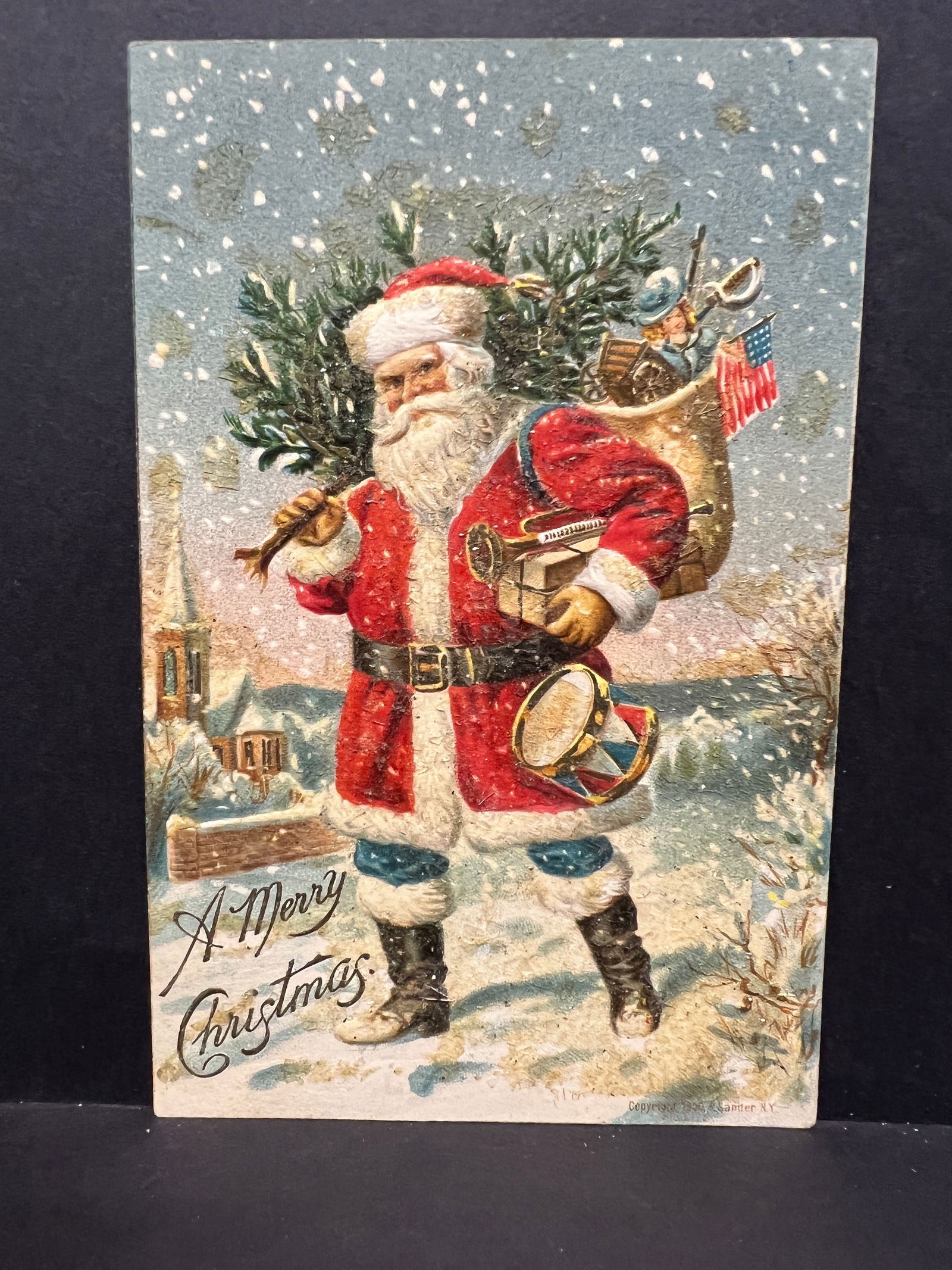 Santa Claus with Toys & Tree Textured Embossed Christmas Postcard, J.I. Austen Co., USA 1906.: An undivided back, illustrated postcard featuring Santa Claus. The front depicts Santa Claus dressed in a traditional red suit with white trim, black belt, and boots, standing in a snowy landscape. He