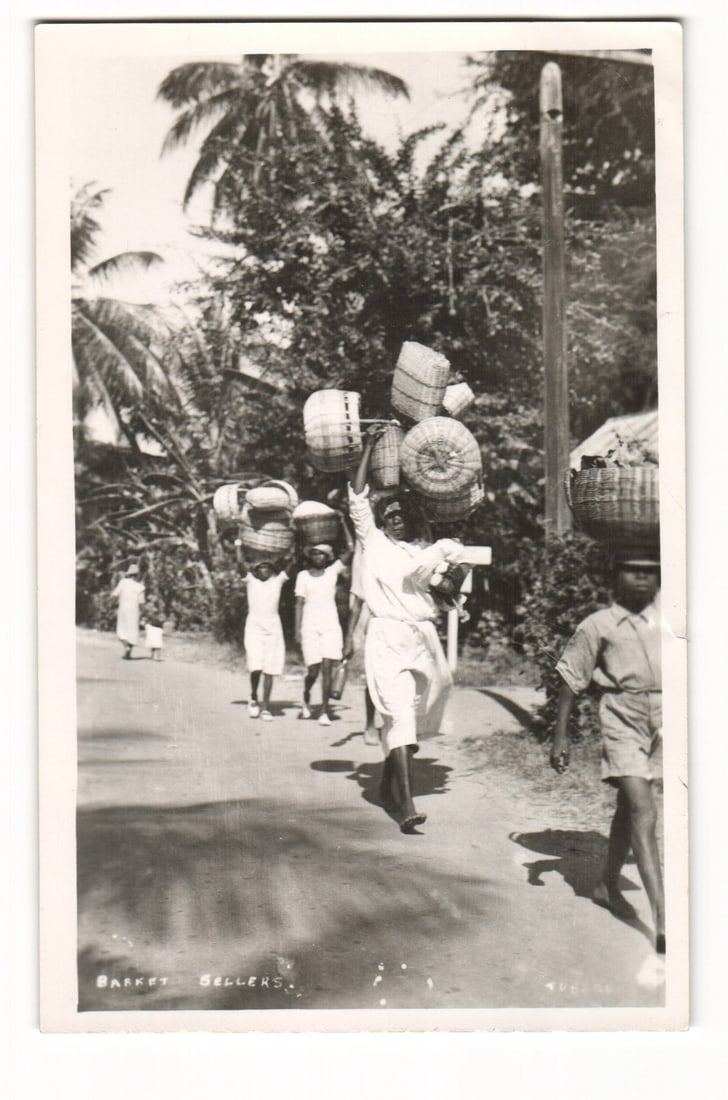 Real Photo Postcard of Basket Sellers on a Tropical Road, Tobago, Early 20th Century Scene: A vertically oriented black and white Real Photo Postcard. The image features a group of individuals identified by printed text at the bottom as "BASKET SELLERS." walking along an unpaved road in a tr