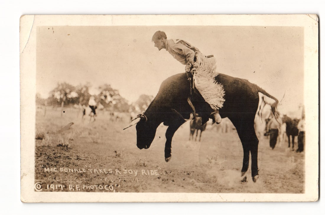 Mac Donald Takes A Joy Ride Bull Riding, U.F. Photo Co. 1917, Vale Oregon 1921 Postcard: A sepia-toned photograph captures a rider, identified by printed text as "MAC DONALD," in profile, engaged in bull riding. The rider, wearing fringed chaps and a light-colored shirt, leans forward as