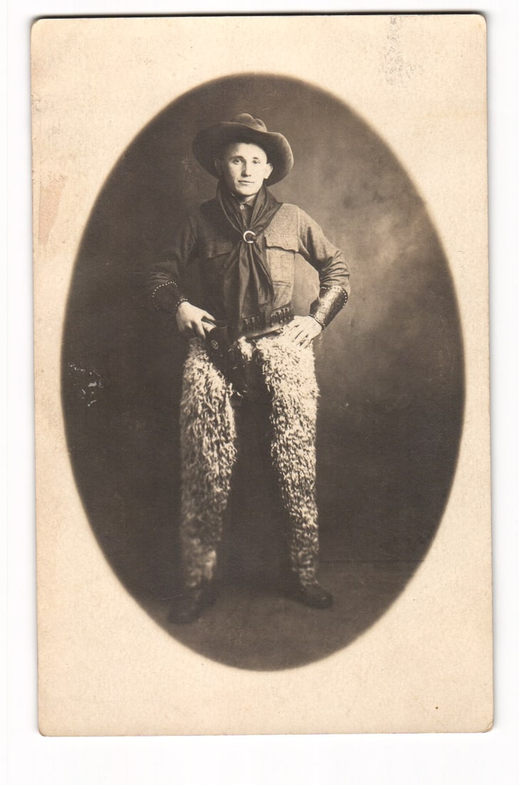 RPPC Studio Portrait of Young Man in Cowboy Attire, Woolly Chaps, Revolver, Early 20th C postcard: A sepia-toned, full-length photographic studio portrait of a young man, presented in an oval vignette. The subject stands facing forward, his right hand resting on a holstered revolver at his hip and