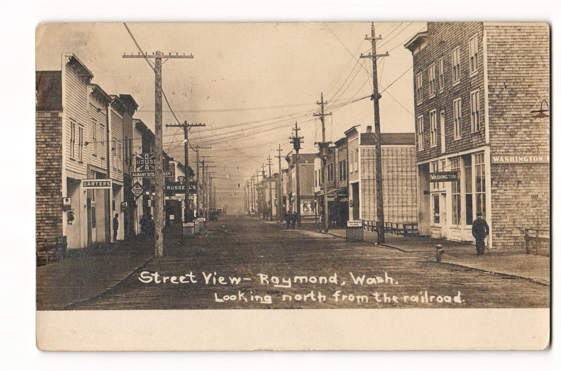 Historic Street View of Raymond, Washington Looking North from Railroad, 1910 RPPC Postcard: A sepia-toned real photo postcard. The image depicts a street scene in Raymond, Washington, looking north from the railroad, as stated by the printed caption. The unpaved street is lined on both sides