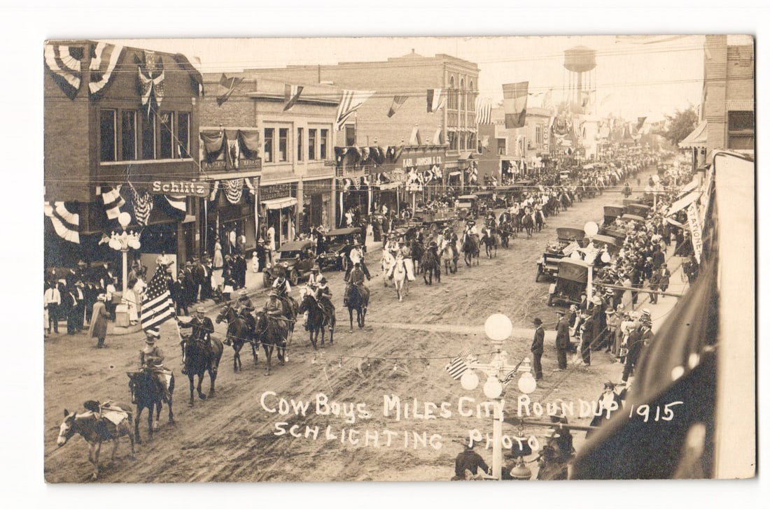 Cowboys Miles City Roundup 1915 Parade, Schlichting Photo, American Flags, Real Photo Postcard: A sepia-toned Real Photo Postcard presenting a high-angle, street-level perspective of a parade. The procession, featuring numerous cowboys on horseback—some carrying American flags—and early auto